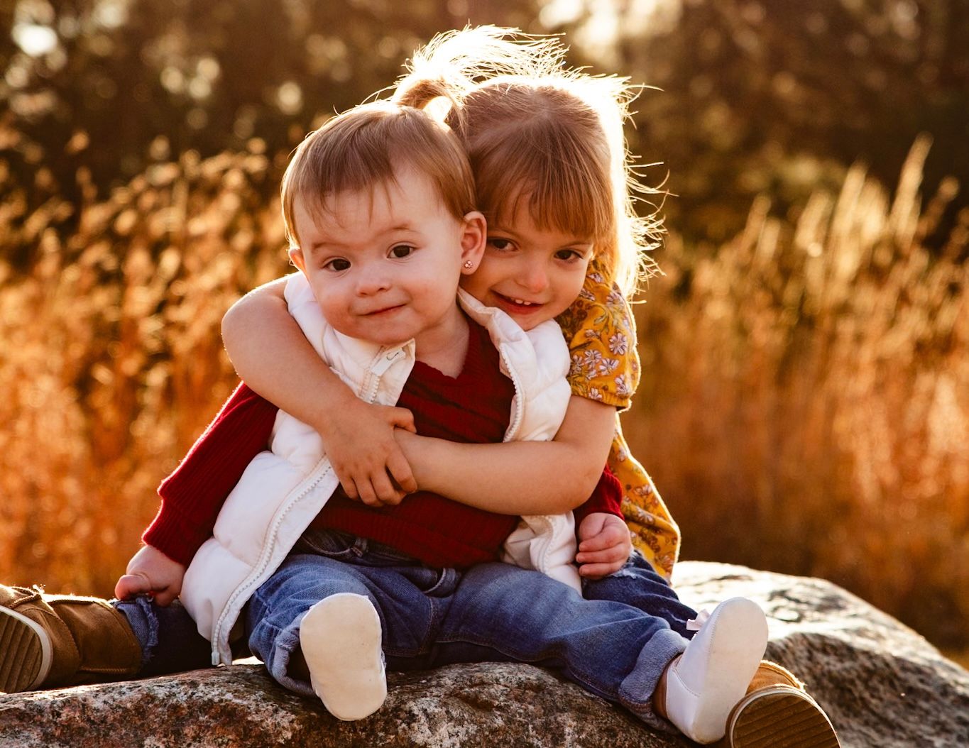 Two young children hugging and sitting on a rock in warm sunset light with golden grasses in background.