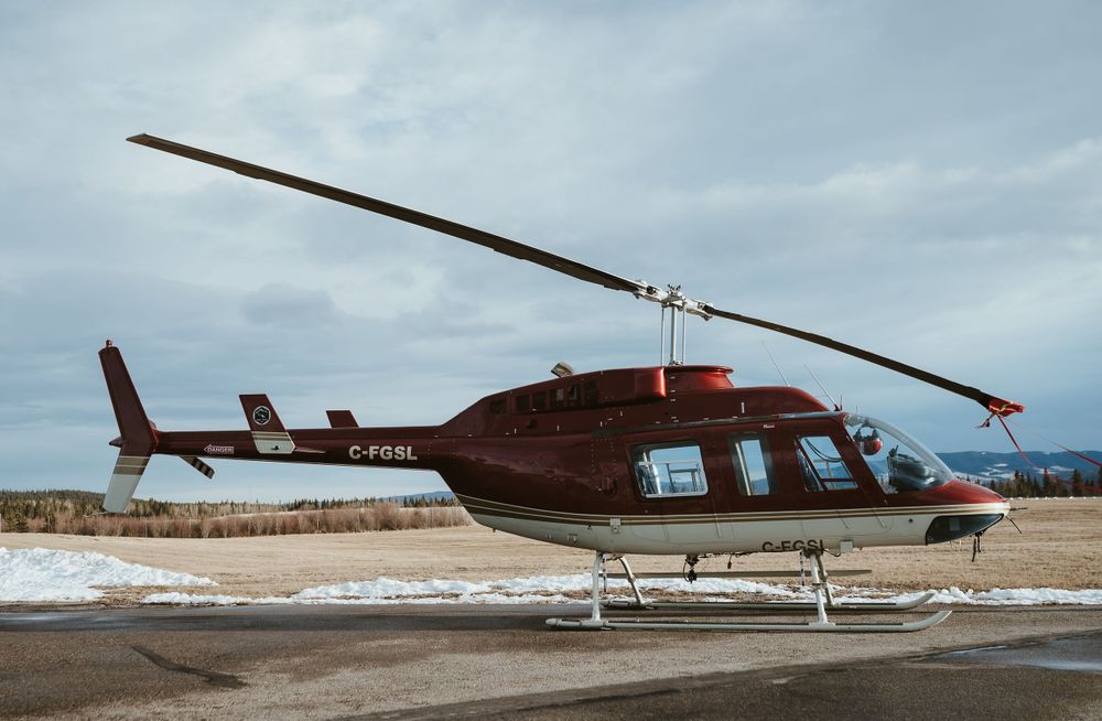 A red and white Bell 206 helicopter parked on snowy ground against cloudy sky.