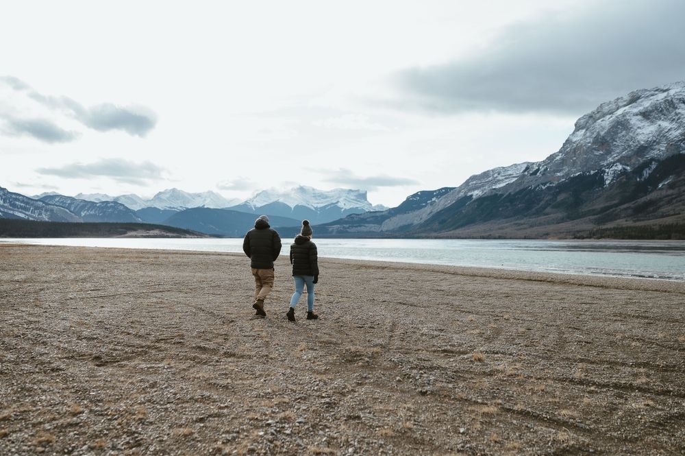Two people walking along a rocky shoreline with mountains and a lake in the background.