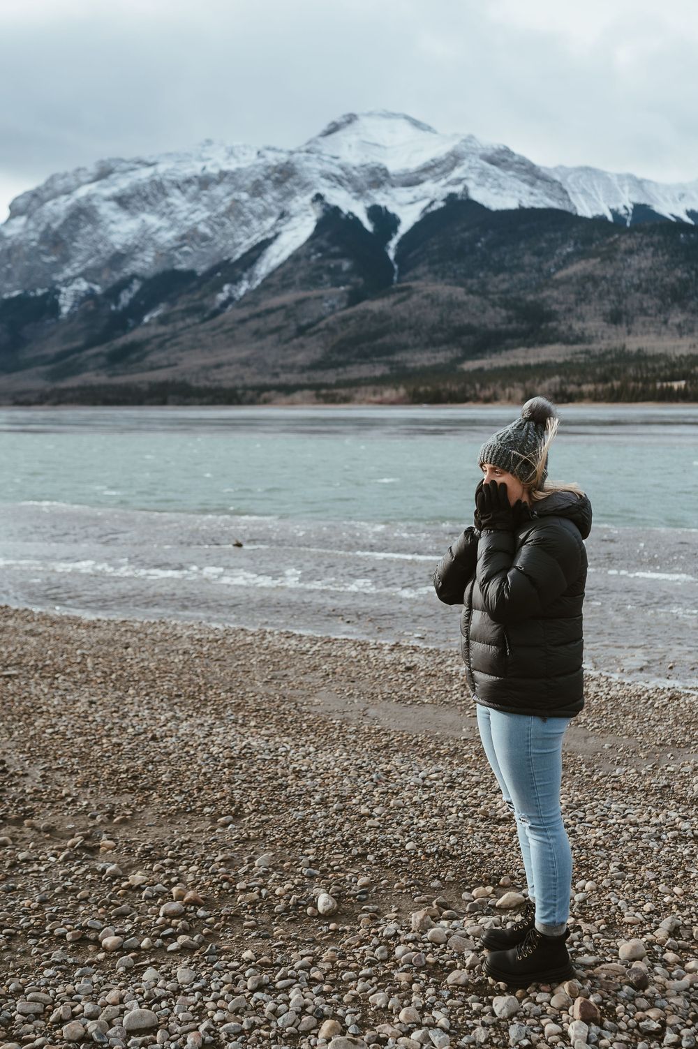 Person in winter clothing taking photos by a frozen lake with snowy mountains in the distance.