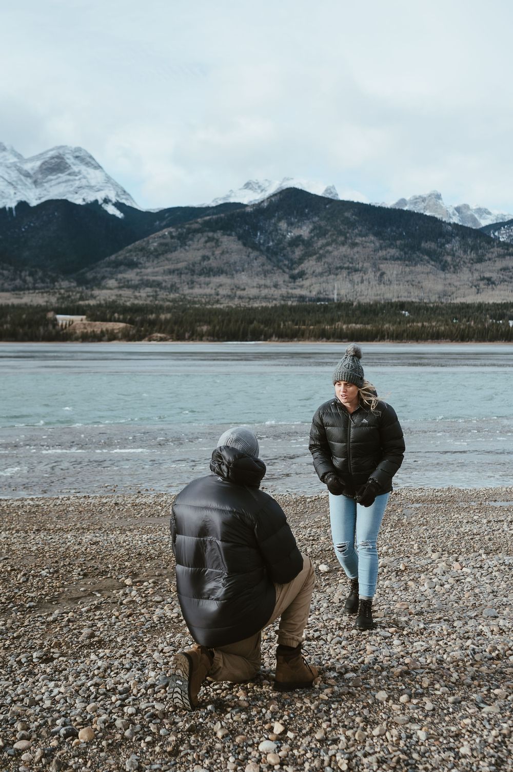 Two people on a rocky shore with snow-capped mountains and frozen lake in the background.