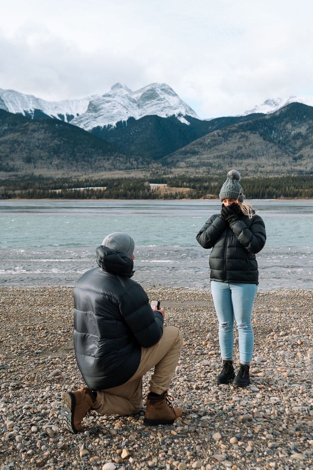 Two individuals on a rocky beach photographing snow-covered mountains beside a frozen lake.