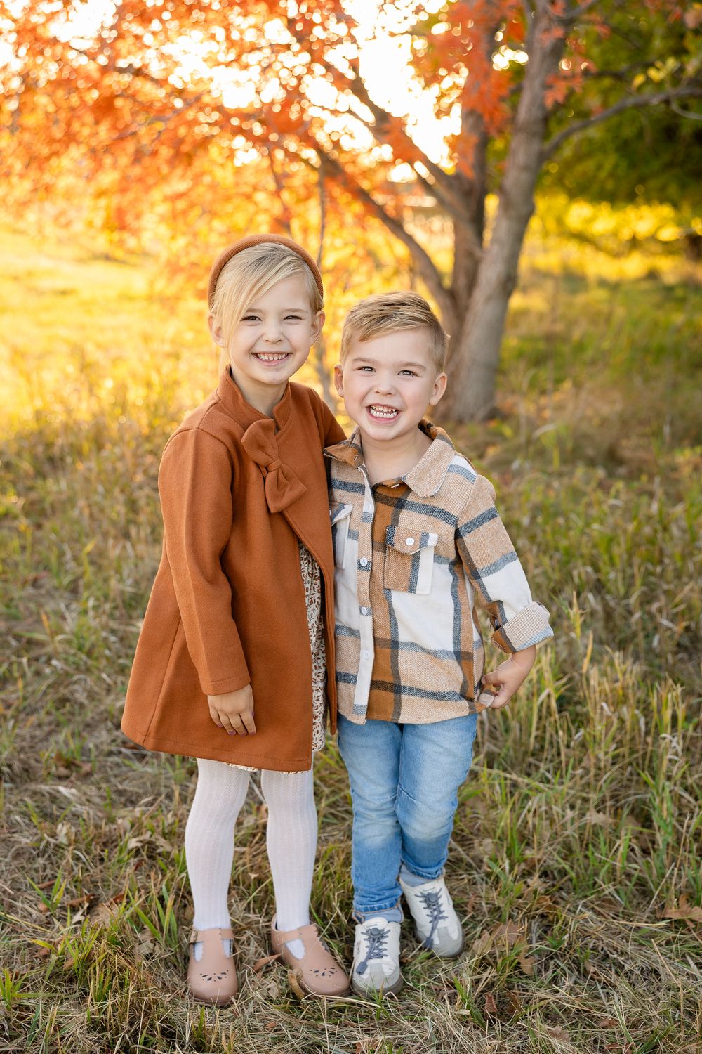 Two children pose together and smile brightly in autumn attire during golden hour.