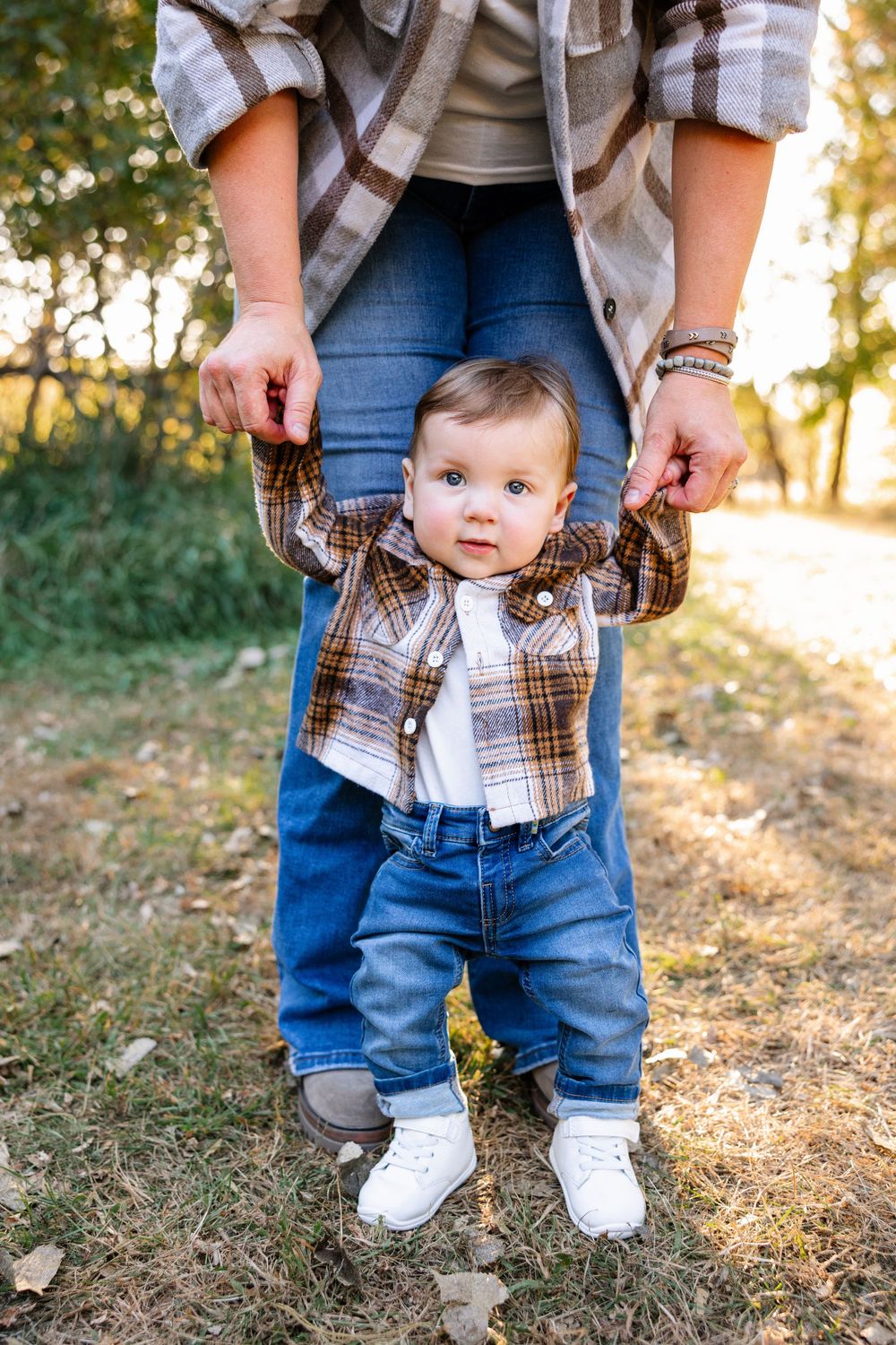 Small child in plaid shirt and jeans being held up while learning to stand outdoors.