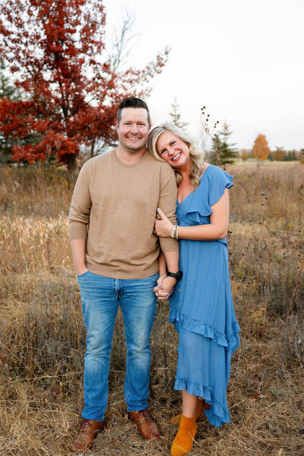 Couple standing together in a fall field with vibrant red foliage in the background.