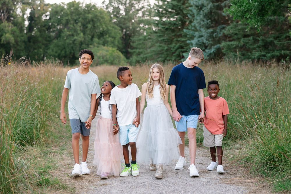 A group of young people in casual attire walk together on a dirt path through tall grass on a sunny day.