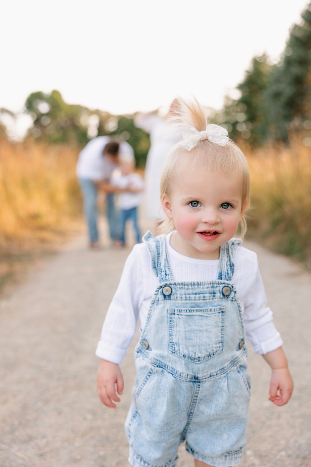 Small child in denim overalls standing on dirt path with blurred figures in background.