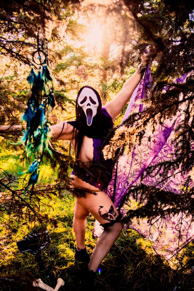 Figure in Ghostface mask poses dramatically with purple and blue decorations in sunlit forest.