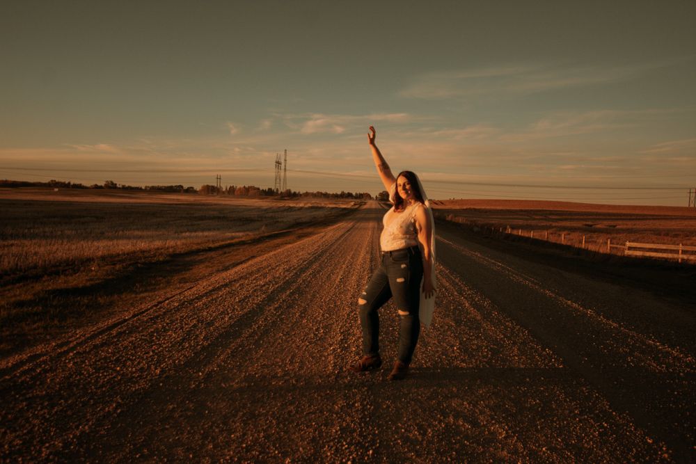 Figure stands joyfully on a rural dirt road during golden hour with arms raised.