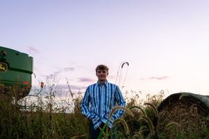 Young man in plaid shirt standing in a field at dusk near a green John Deere tractor.