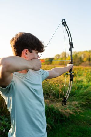 Young man drawing a compound bow outdoors in a field, practicing archery on a sunny day.