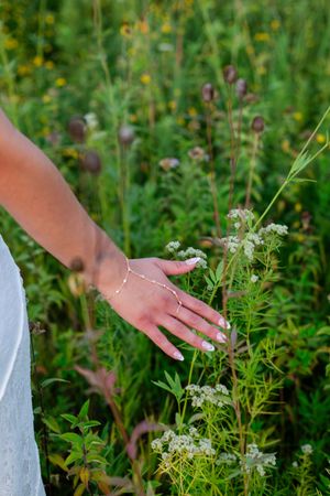 A woman's hand with a gold bracelet and ring gently touching white wildflowers in a lush green meadow.