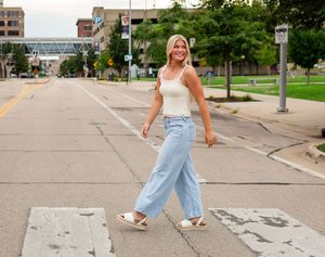 Smiling blonde woman in white tank top and light blue wide-leg jeans walking across a crosswalk in an urban setting.