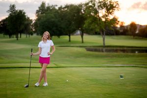 Woman golfer in pink skirt smiling on lush green golf course at sunset holding a driver club.