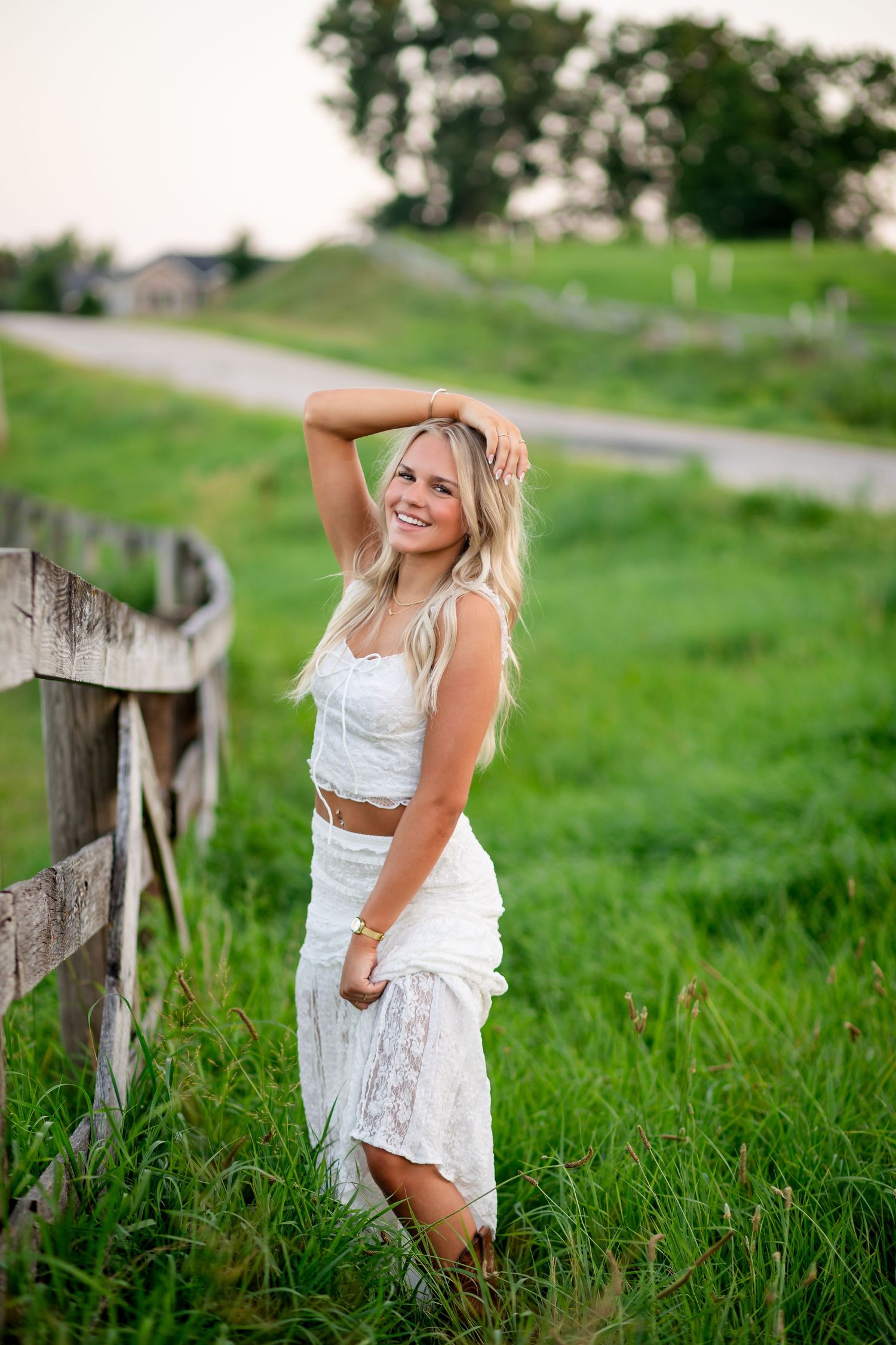 Smiling blonde woman in white lace crop top and skirt posing by a wooden fence in a lush green countryside field.