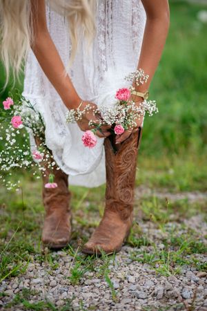 Woman in white lace dress and brown cowboy boots holding pink roses and baby's breath flowers outdoors.