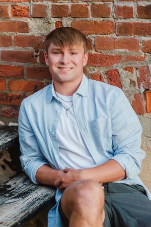 Young man in light blue shirt smiling while seated against a rustic brick wall outdoors.