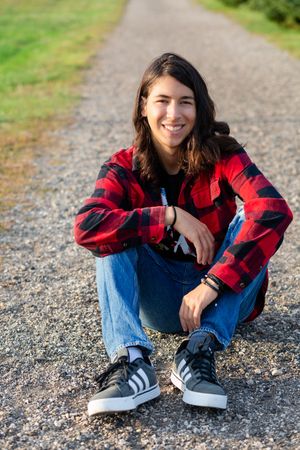 Smiling teen with long hair sits on gravel path wearing red plaid shirt, jeans, and Adidas sneakers.