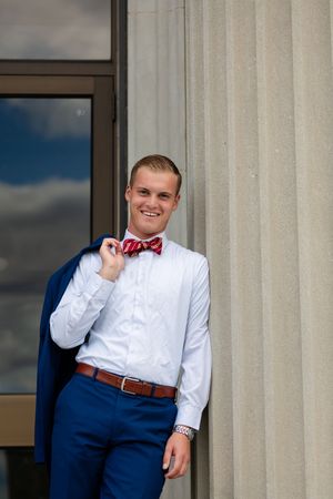 Smiling young man in white shirt, red bow tie, blue pants, holding navy blazer over shoulder by stone columns.