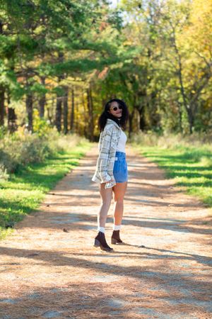 Young woman in plaid shirt, denim shorts, and boots posing on a scenic forest path in autumn.