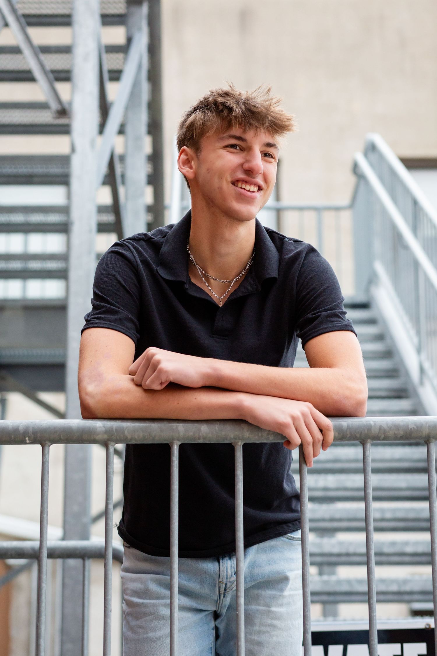 Young man in black polo shirt leaning on metal railing, smiling outdoors near industrial stairs.