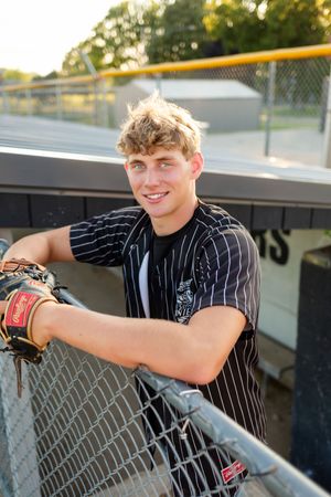 Young baseball player in pinstripe jersey smiles leaning on dugout fence with Rawlings glove at outdoor ballpark.