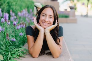 Smiling young woman resting chin on hands outdoors near purple flowers in a garden setting.