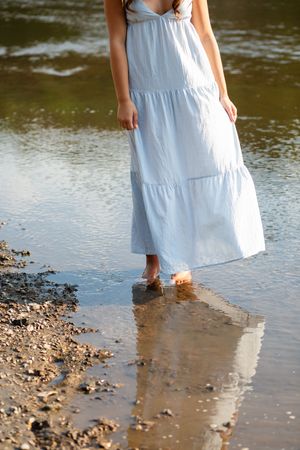 Woman in white maxi dress wading barefoot in shallow water near a rocky shoreline.