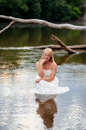 Smiling blonde woman in white dress sitting in a calm river with fallen logs and green trees in the background.