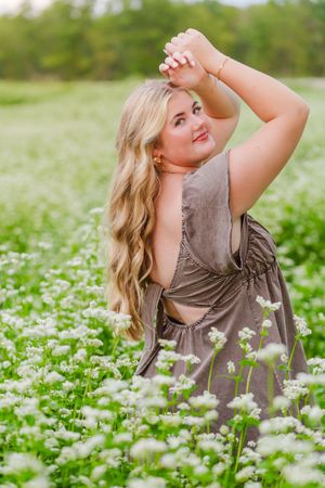 Blonde woman in gray backless dress posing in a field of white wildflowers with green trees in background.