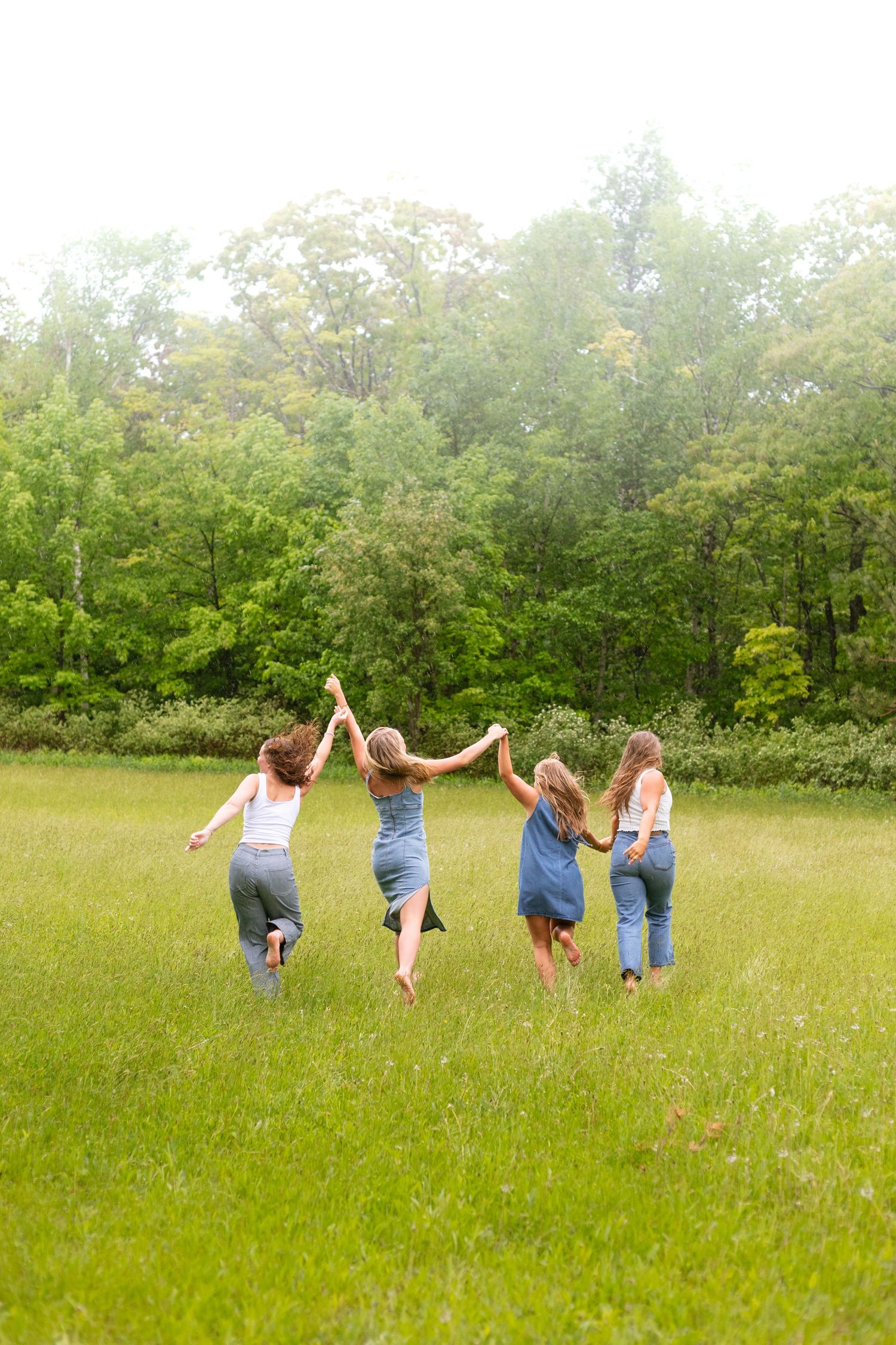 Four girls holding hands run through a lush green meadow toward a forest on a cloudy day.