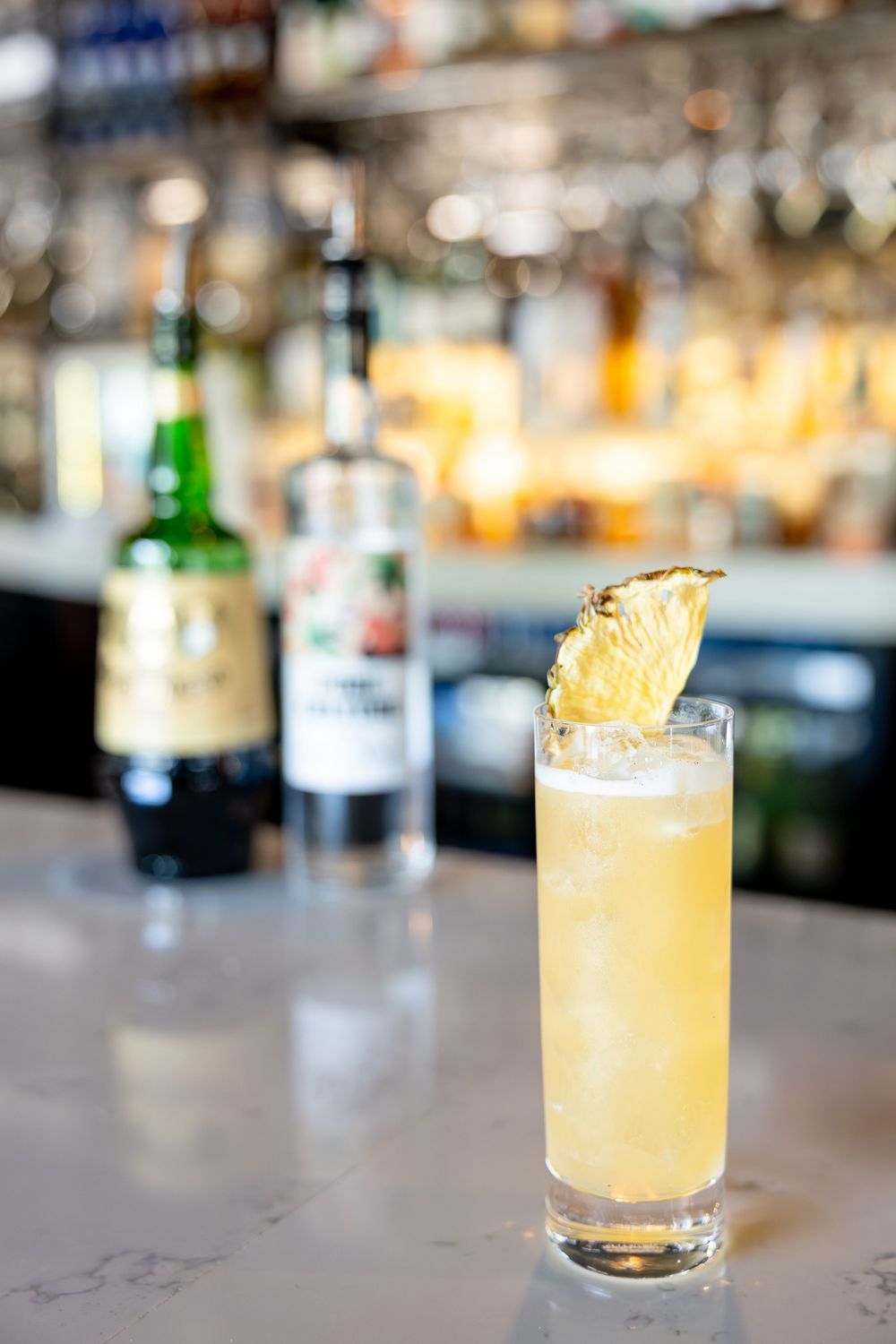 Pineapple-garnished tall cocktail on a marble bar with liquor bottles in the background.