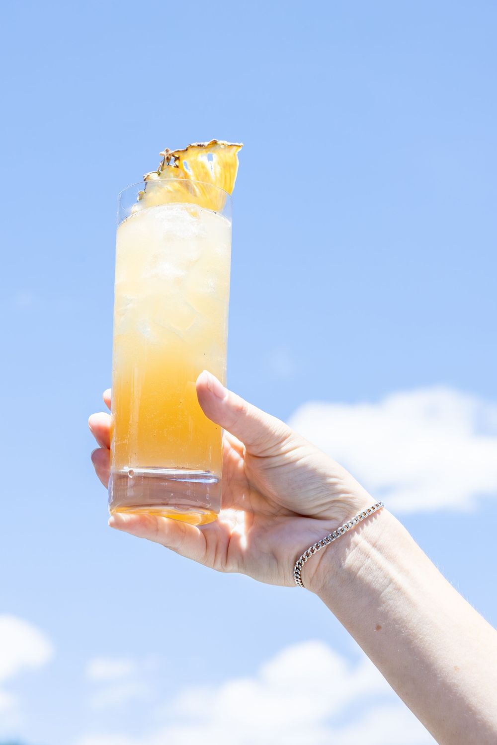 Hand holding a tall glass of icy pineapple cocktail garnished with a fresh pineapple slice against a blue sky.
