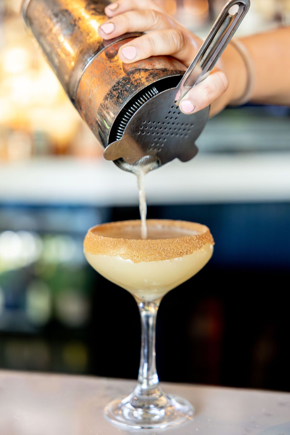 Bartender straining a cocktail into a sugar-rimmed coupe glass at a bar.