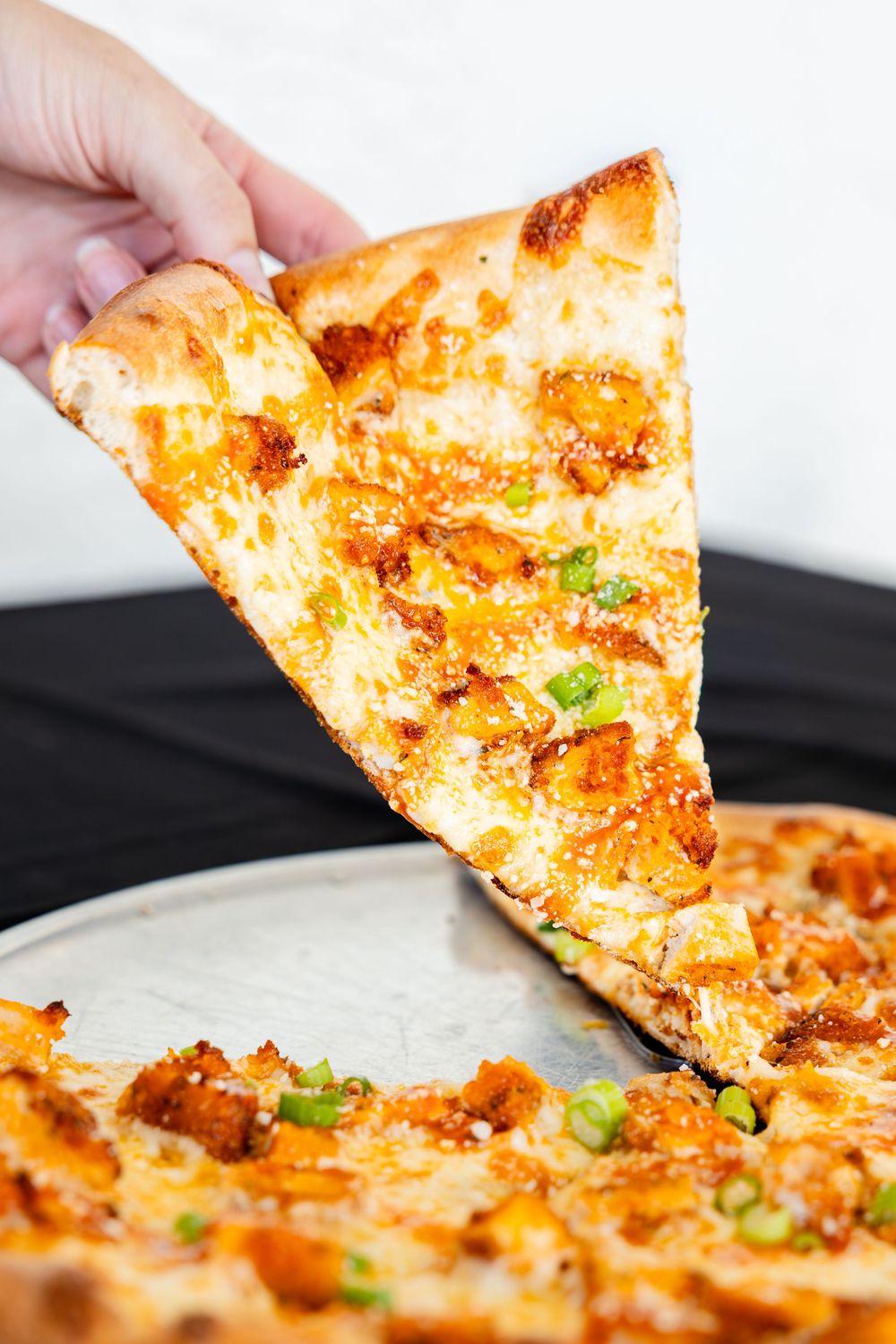 Hand lifting a cheesy slice of buffalo chicken pizza topped with green onions from a metal pan.