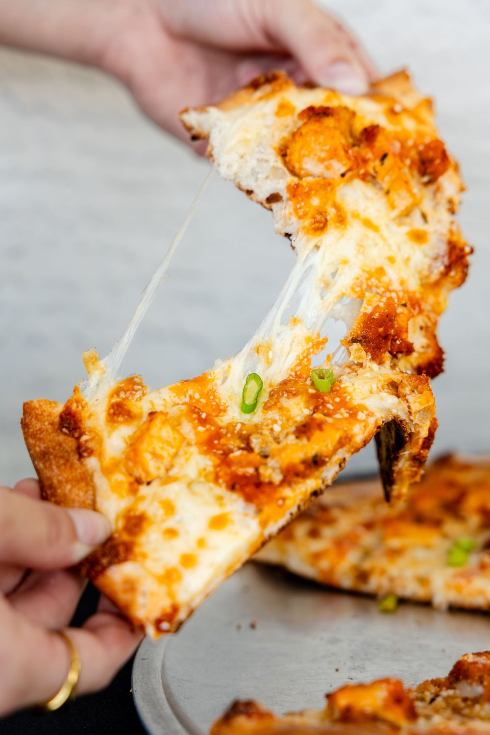 Hands pulling apart a cheesy buffalo chicken pizza slice with melted cheese stretching between pieces.