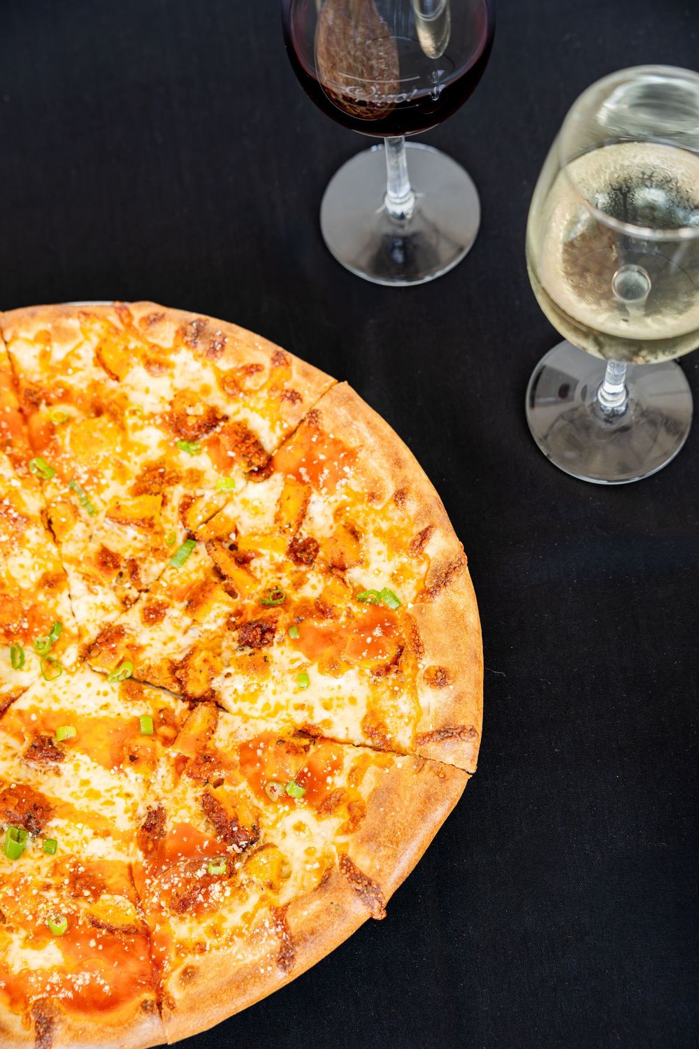 Whole buffalo chicken pizza topped with melted cheese and green onions served beside two glasses of wine.