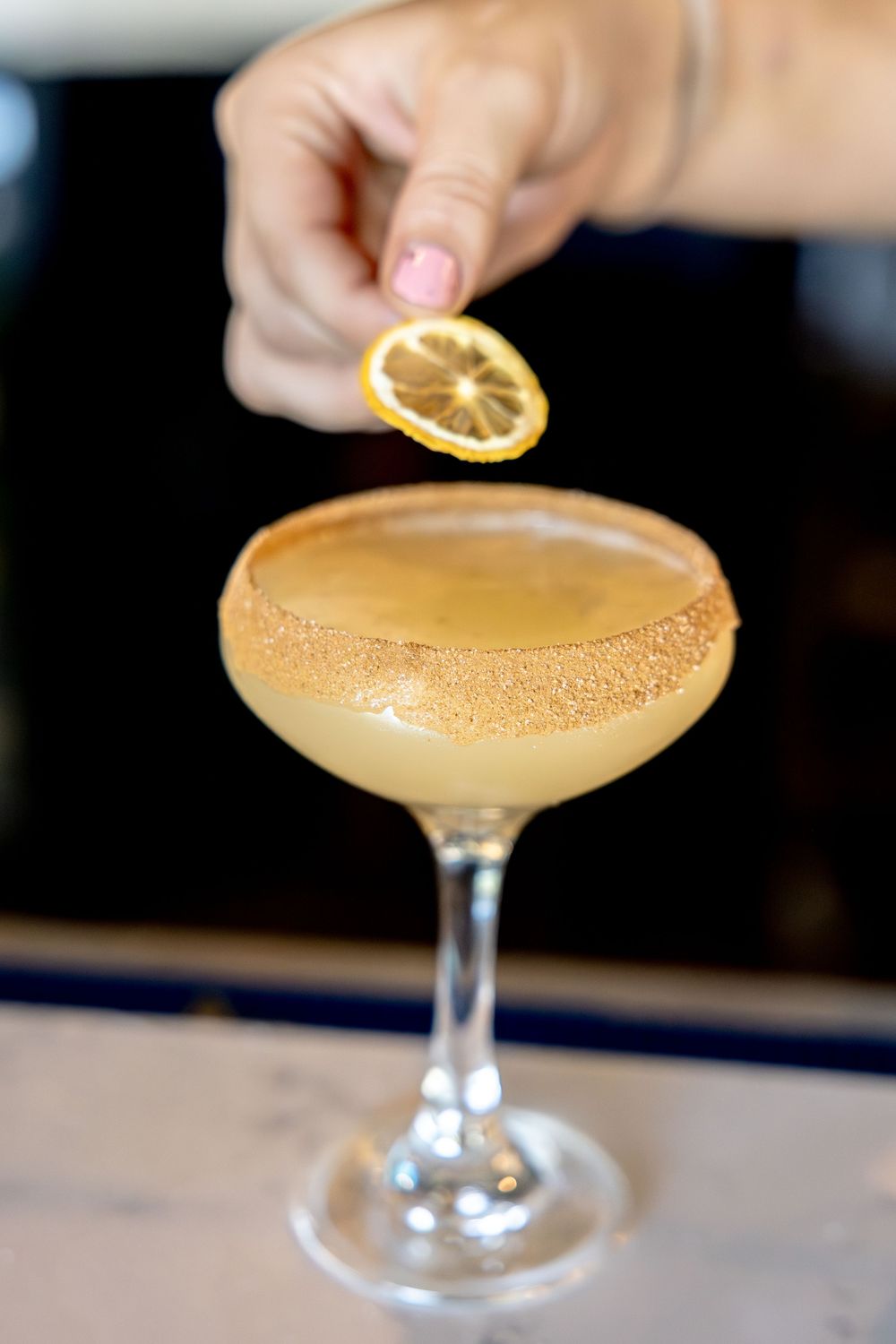 Bartender garnishing a sugar-rimmed coupe cocktail with a dried lemon slice.