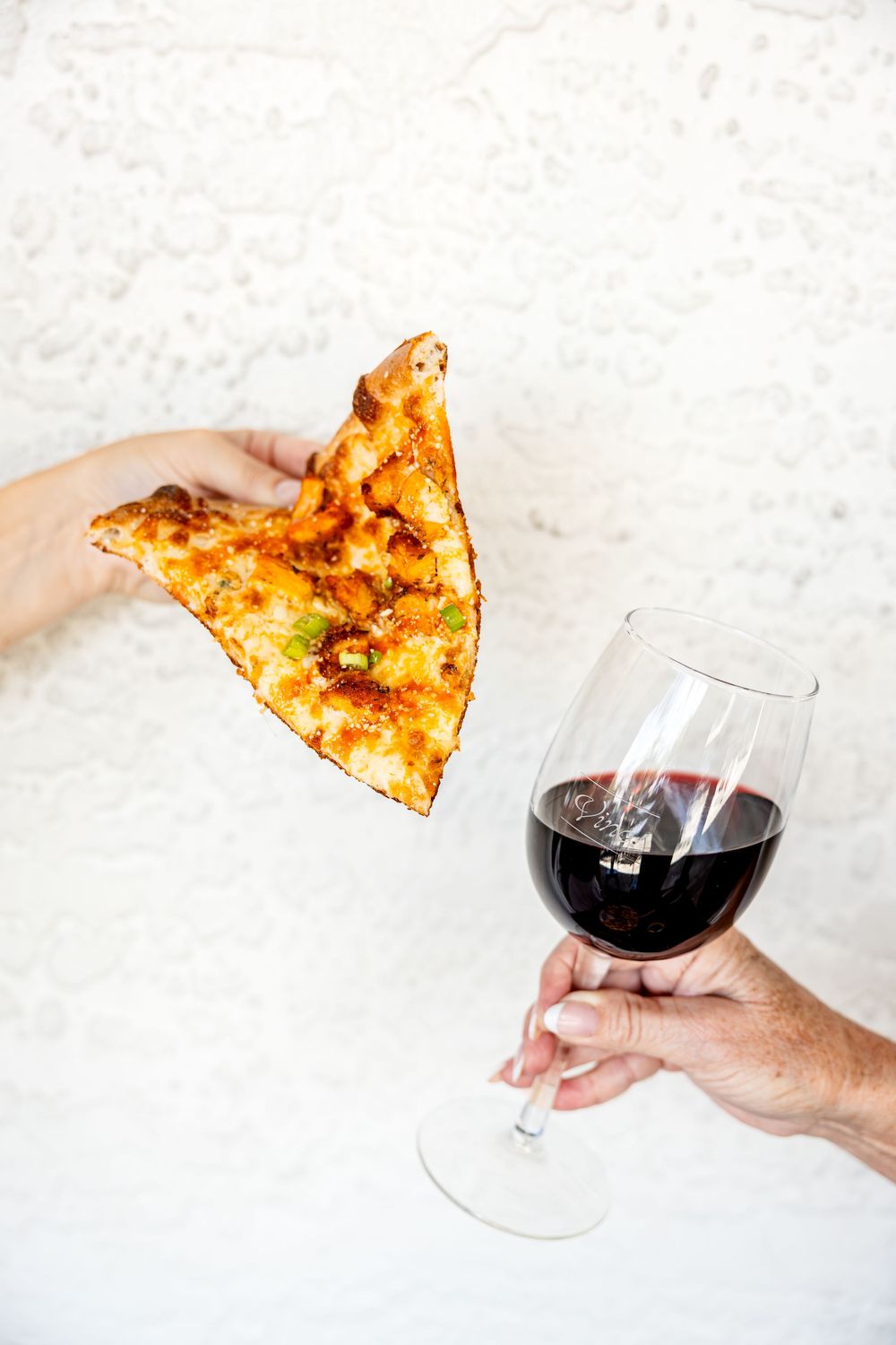 Hands holding a slice of buffalo chicken pizza and a glass of red wine against a white textured background.