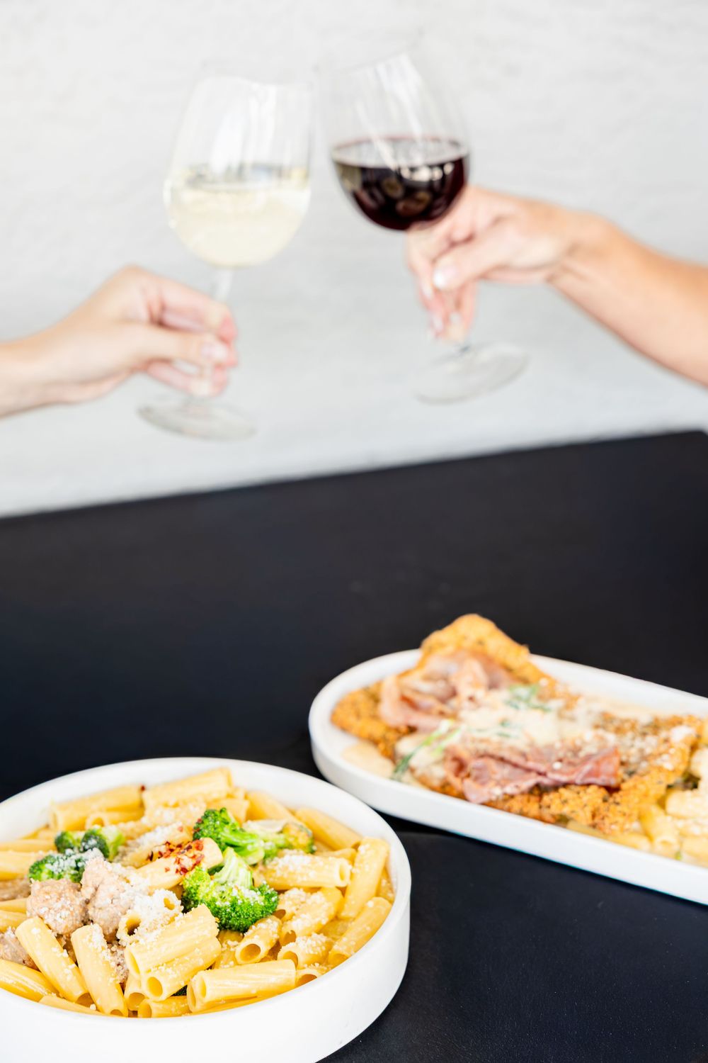 Two hands toasting wine glasses over a table with penne pasta and breaded chicken dishes.