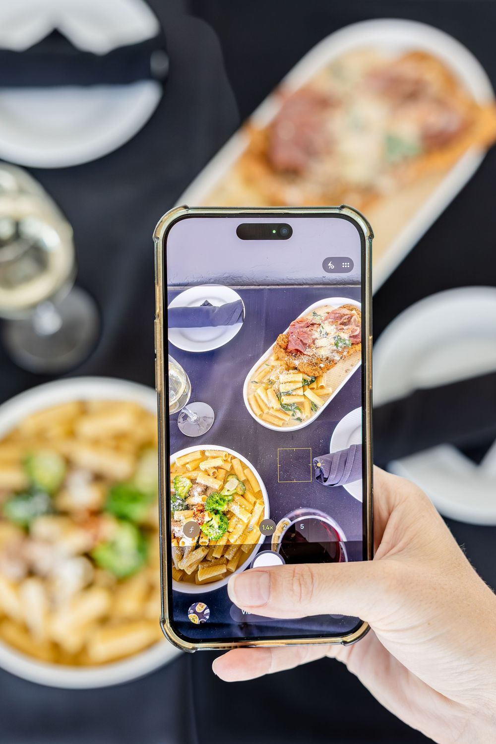 Person photographing Italian food spread including pasta, breaded chicken, and wine on a smartphone.