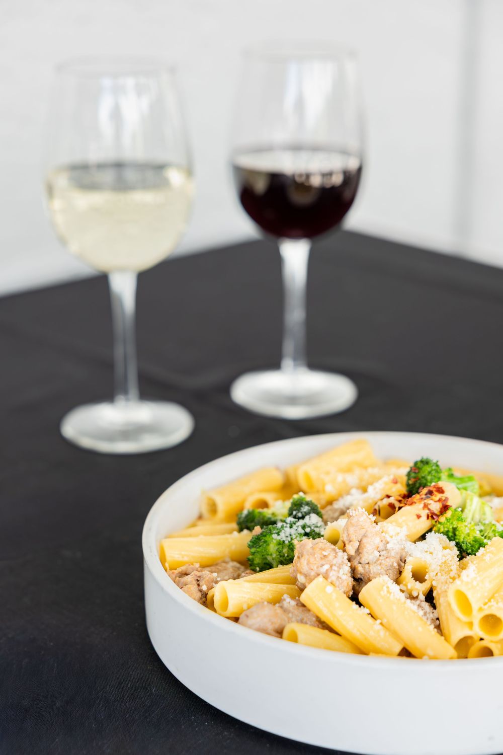 Bowl of penne pasta with broccoli and chicken in creamy sauce alongside red and white wine glasses.