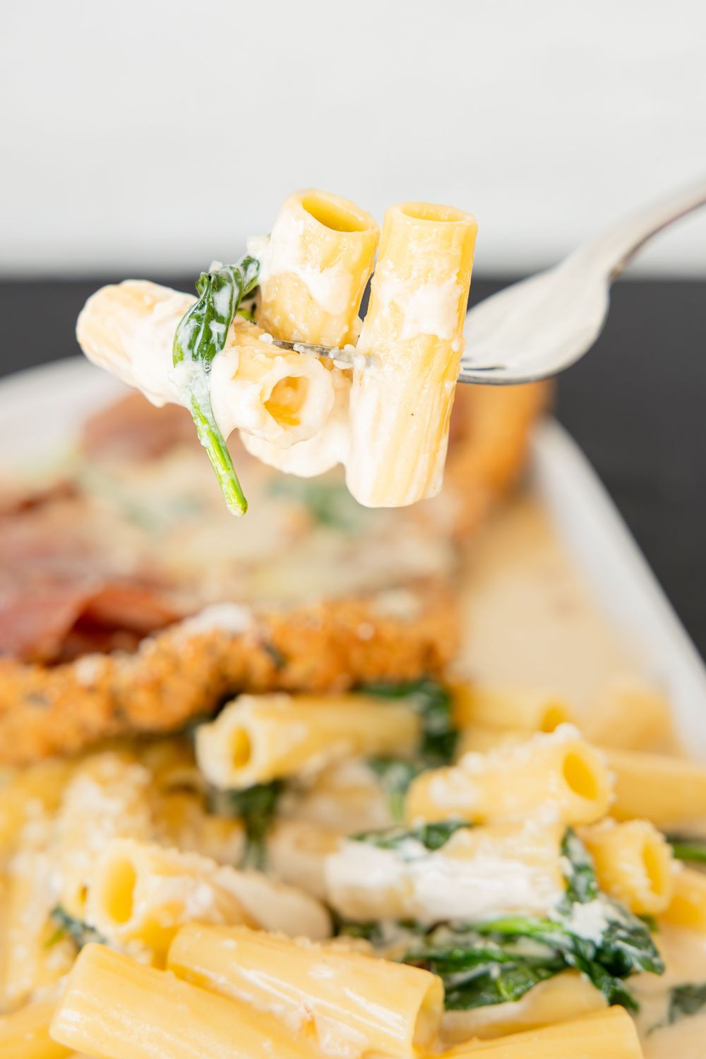 Fork holding rigatoni pasta with spinach in creamy sauce above a dish of breaded chicken and pasta.