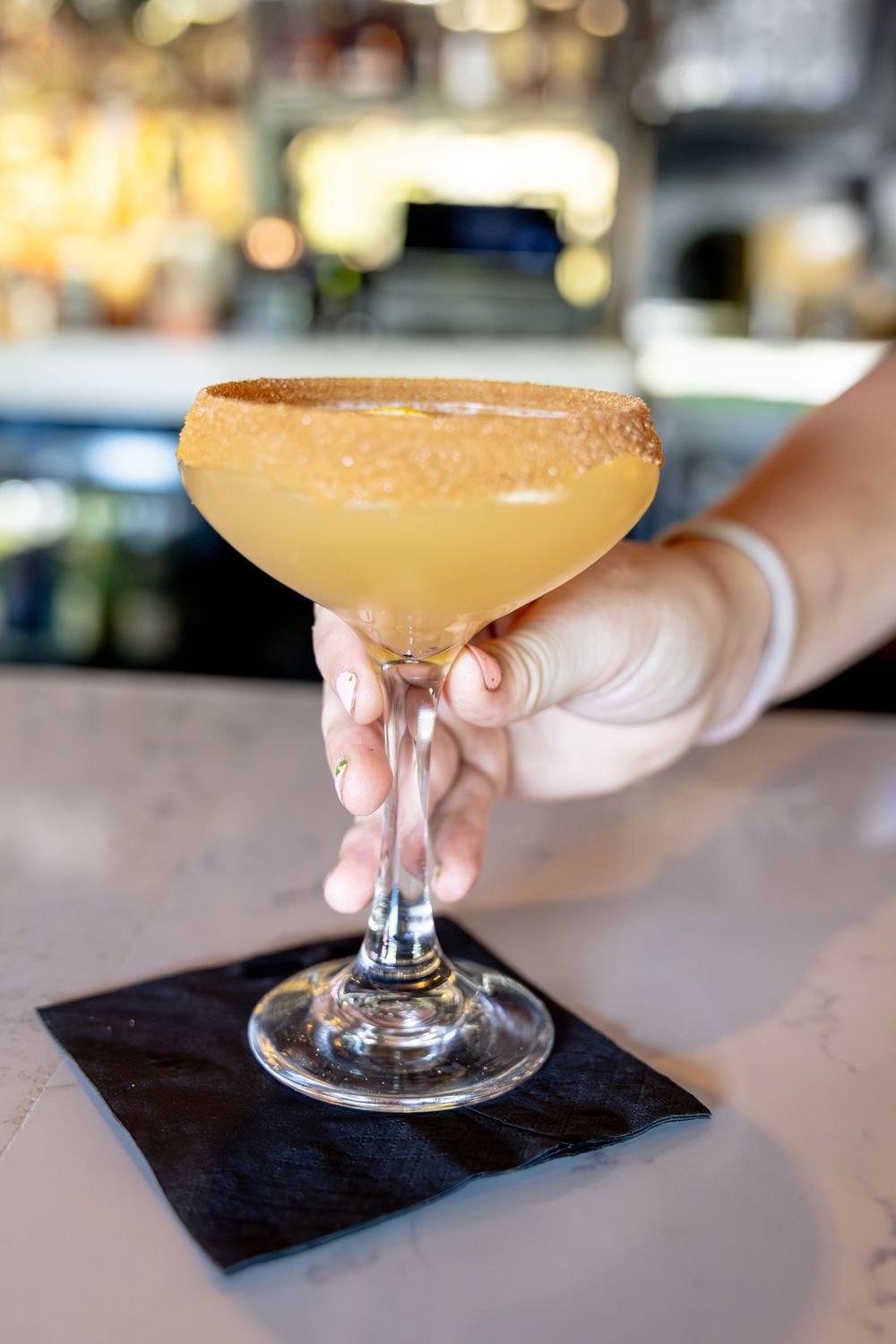 Bartender presenting a sugar-rimmed yellow cocktail in a coupe glass on a napkin.