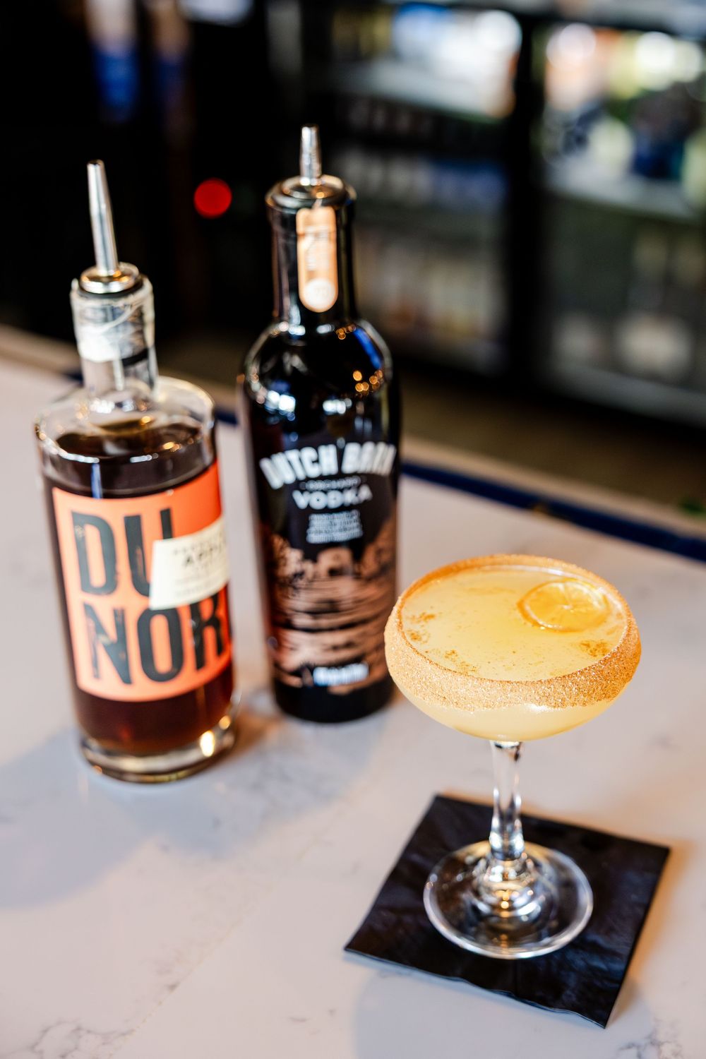 Golden cocktail in a sugar-rimmed coupe glass beside Du Nord and Hatch Bank vodka bottles.