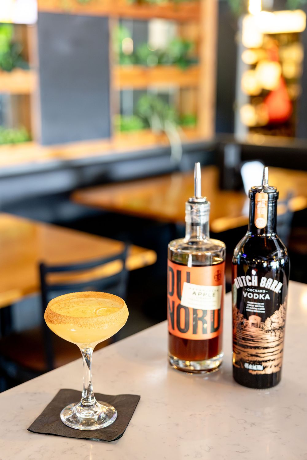 Yellow cocktail in a coupe glass beside Du Nord and Hatch Bank vodka bottles on a bar.