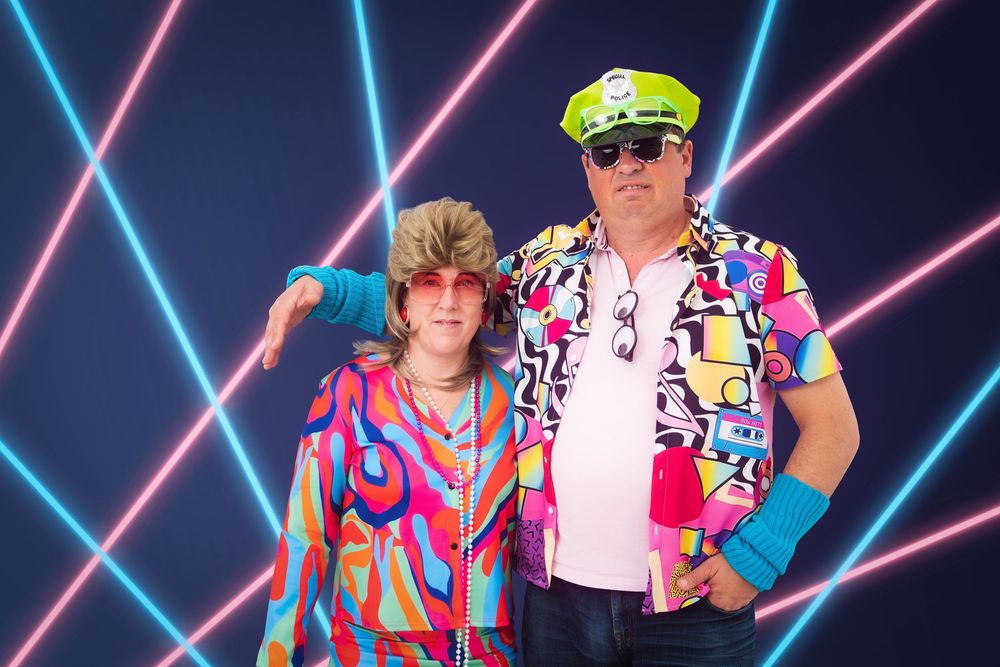 a couple dressed in 80's clothes standing awkwardly beside each other, taken in front of an 80's neon laser background.