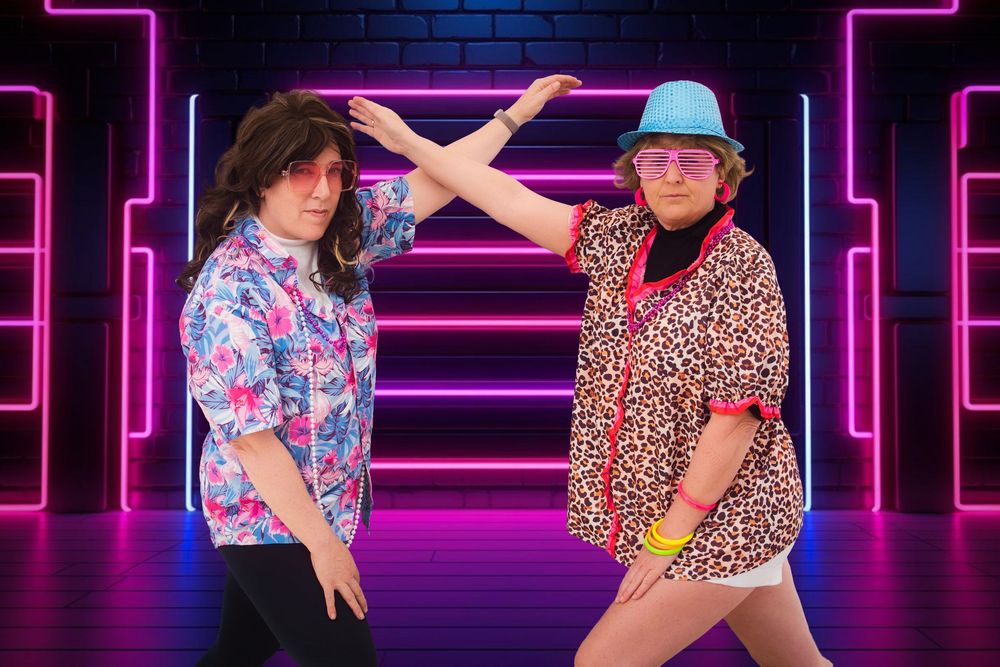 Awkward photo of two women dressed in 80's clothes doing a funny pose in front of a neon background