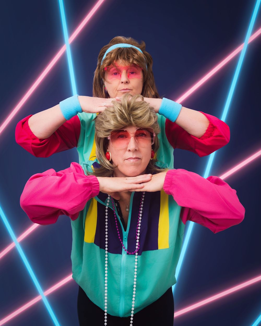 Awkward photo of two women dressed in 80's clothes doing a funny pose of stacked heads, in front of a neon laser background