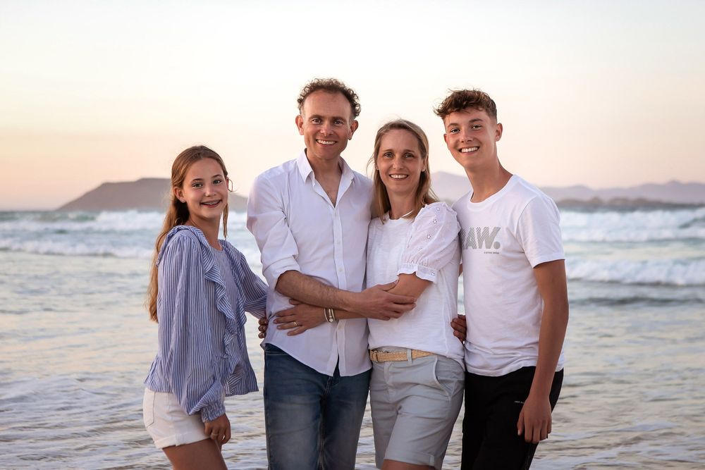 Family of four smiling together on a beach at sunset, with ocean waves and mountains in the background.